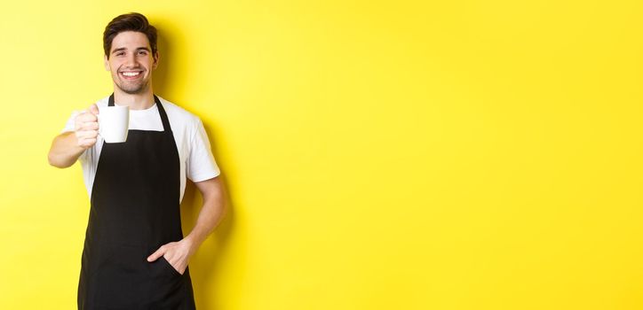 Handsome barista in black apron giving you cup of coffee and smiling, standing over yellow background.