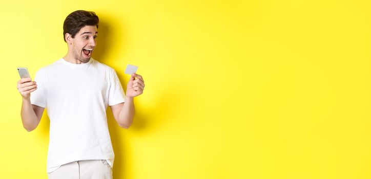 Surprised guy holding smartphone and credit card, online shopping on black friday, standing over yellow background.