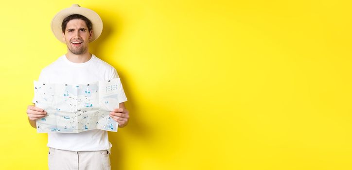 Travelling, vacation and tourism concept. Puzzled guy tourist cant understand map, looking confused at camera, standing against yellow background.