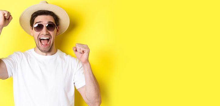 Concept of tourism and holidays. Happy male tourist celebrating his vacation, raising hands up and shouting for joy, wearing sunglasses with straw hat.