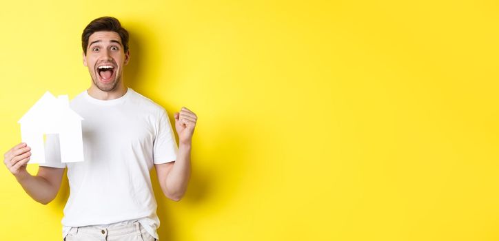 Real estate concept. Excited man holding paper house model and celebrating, standing happy over yellow background.