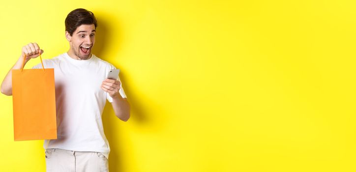 Concept of discounts, online banking and cashback. Surprised man showing shopping bag and looking happy at mobile screen, standing against yellow background.