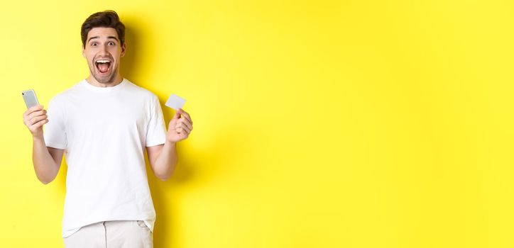 Surprised guy holding smartphone and credit card, online shopping on black friday, standing over yellow background.