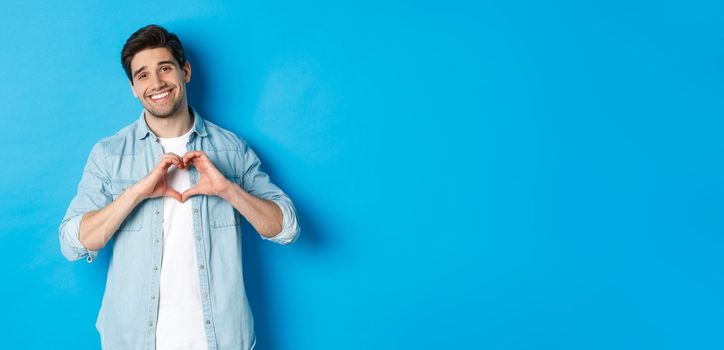 Handsome man smiling, showing heart gesture and looking at camera, saying I love you, standing against blue background.