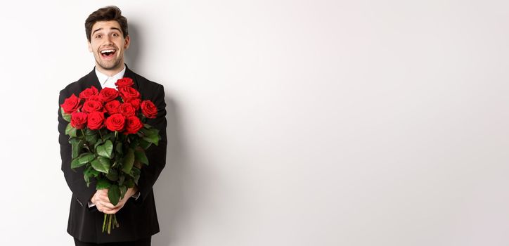 Image of handsome boyfriend in black suit, holding bouquet of red roses and smiling, being on a date, standing over white background.