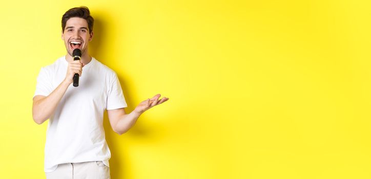 Happy man entertainer performing, talking in microphone, making speech or stand-up show, standing over yellow background.