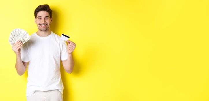 Young man withdraw money from credit card, smiling pleased, standing over yellow background.