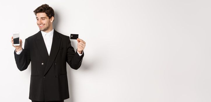 Handsome successful businessman, looking at smartphone screen and showing credit card, standing in black suit against white background.