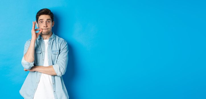 Adult man zipping mouth, promise keep secret, making a seal on lips and standing over blue background.