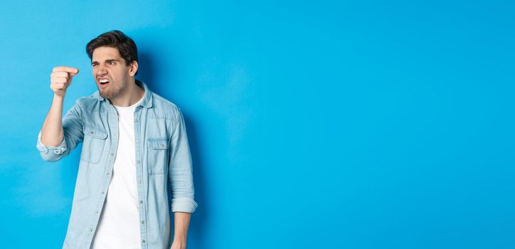 Confused guy holding something tiny in fingers and looking at it puzzled, standing over blue background.