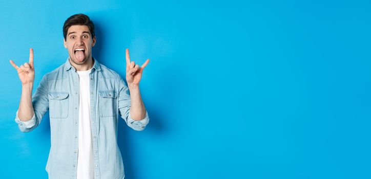 Cool guy having fun, enjoying concert, showing rock sign and tongue, standing over blue background.