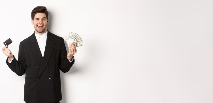 Image of confident businessman in black suit, smiling pleased and winking, holding money and credit card, standing against white background.