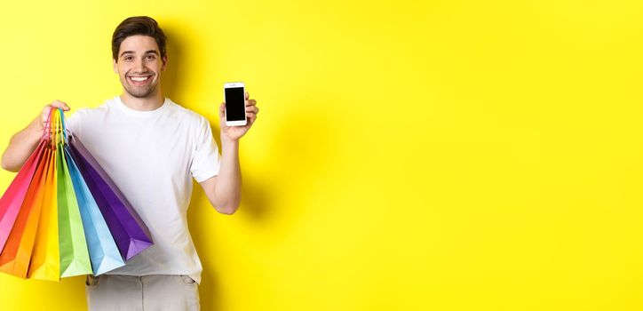 Young man holding shopping bags and showing mobile phone screen, money application, standing over yellow background.