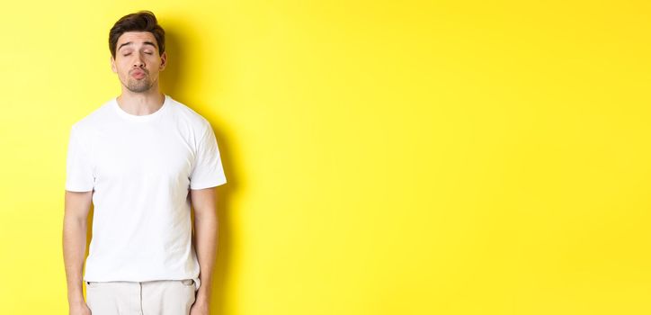 Image of lovely man close eyes and pucker lips, waiting for kiss, standing in white clothes against yellow background. Copy space
