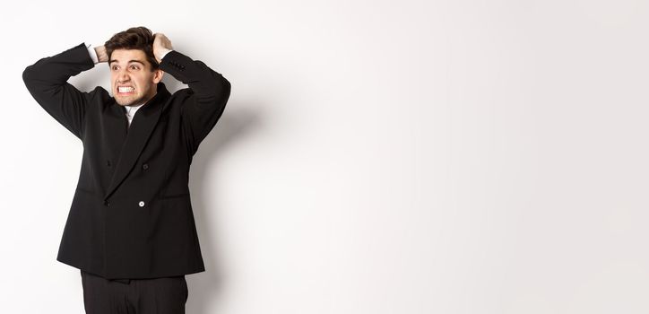 Image of frustrated and angry businessman in black suit, ripping hair on head and grimacing mad, looking left at disaster, standing tensed against white background.