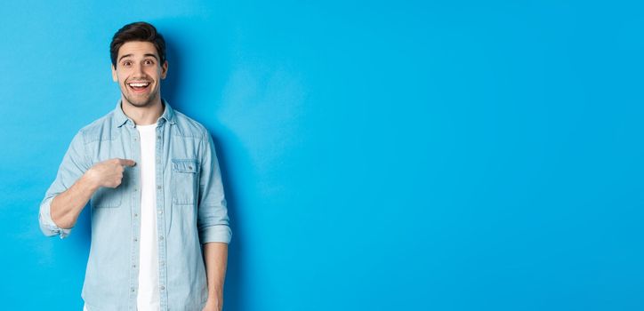 Happy and surprised man pointing at himself, smiling pleased, standing against blue background.