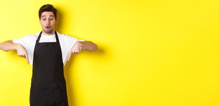 Surprised male barista in black apron checking out promo offer, pointing fingers down at logo banner, standing over yellow background.