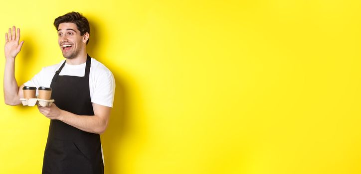 Friendly waiter in cafe waving hand at customer, holding takeaway coffee oder, standing against yellow background in black apron.