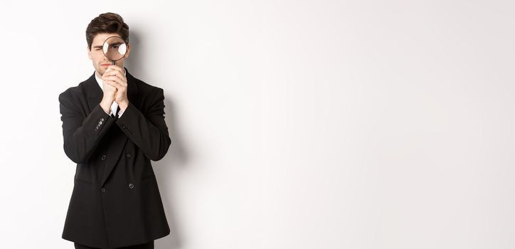 Image of serious businessman in black trendy suit, looking through magnifying glass, looking for employees, standing against white background.