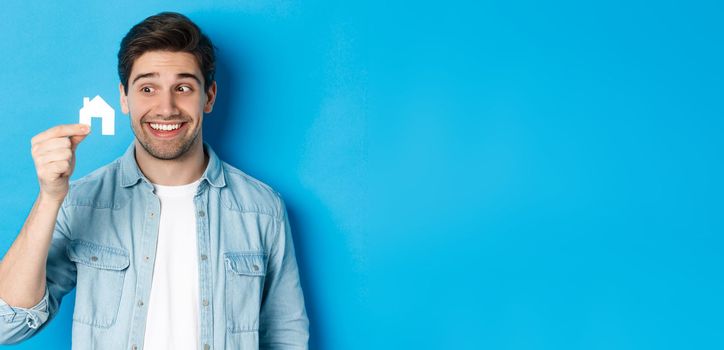 Real estate concept. Excited guy looking at small house model and smiling, renting apartment, standing over blue background.