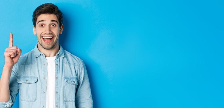 Close-up of handsome bearded guy smiling, showing finger number one, standing over blue background.