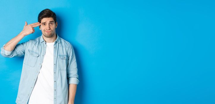 Tired and annoyed adult man making finger gun gesture near head, shooting himself from boredom, standing against blue background.