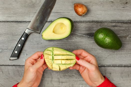 Tabletop view, woman hands holding avocado with cuts, preparing it, removing pulp. Whole avocado, seed and knife on gray wood desk next to it.