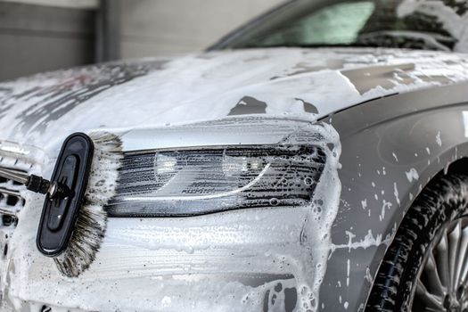 Front light of silver car, covered with shampoo and foam, being washed in manual carwash with brush.