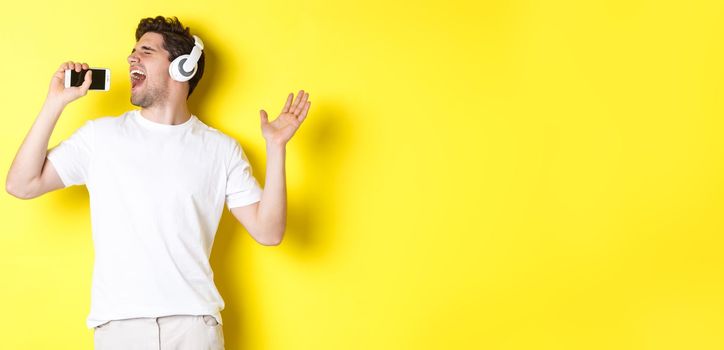 Happy guy playing karaoke app in headphones, singing into smartphone microphone, standing over yellow background.