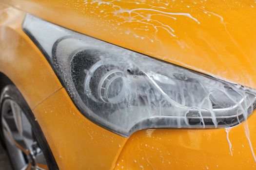 Front light of yellow car washed in carwash, jet water spraying, white shampoo and foam flowing on surface.