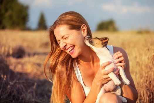 Young woman holding Jack Russell terrier puppy on her hand, trying to pose, but the dog is licking and chewing her ear and hair.