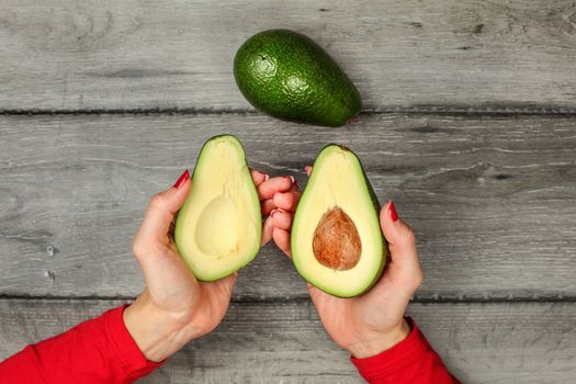 Tabletop view woman's hand holding two avocado halves, seed visible, whole avocado pear next to it.