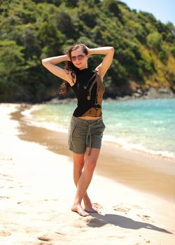 Young woman in black t shirt, shorts and sunglasses, standing on the beach, hands in her hair, sea and tree covered hills in background.