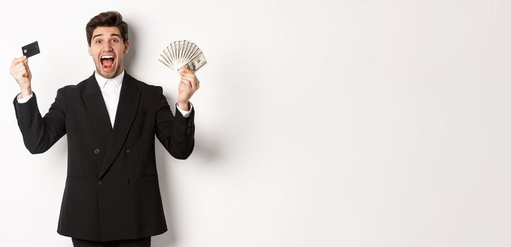 Portrait of handsome businessman in black suit, showing credit card and money, shouting for joy and excitement, standing against white background.