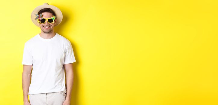 Concept of tourism and vacation. Relaxed smiling man enjoying supper trip, wearing sunglasses and straw hat, yellow background.