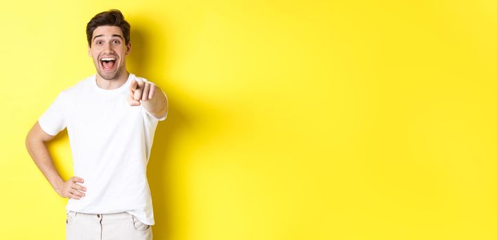 Happy guy pointing finger at camera and laughing, check out something, standing over yellow background.