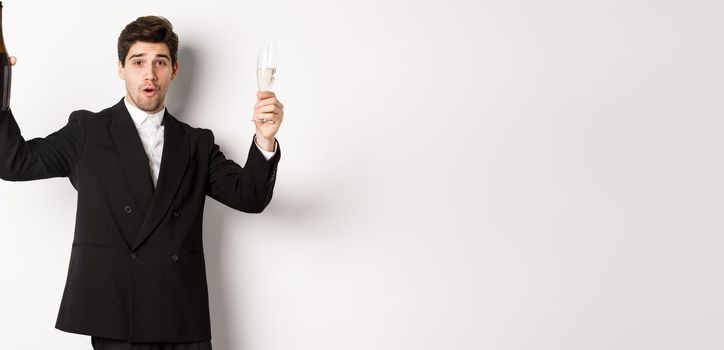 Concept of holidays, party and celebration. Image of handsome man in stylish suit, dancing with bottle of champagne, drinking on new year, standing over white background.