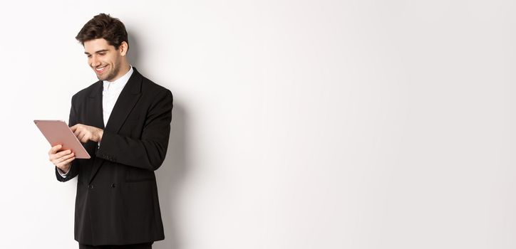 Portrait of attractive man in trendy suit, looking at digital tablet and smiling, shopping online, standing over white background.