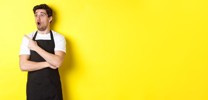 Waiter in black apron checking out promo offer, pointing finger and looking left at your logo, standing over yellow background.