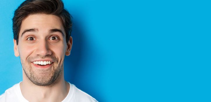 Head shot of handsome male model looking amazed, smiling wondered, standing over blue background. Copy space