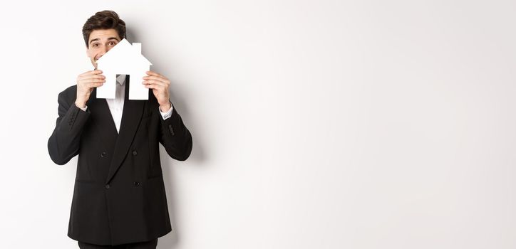 Image of handsome broker in black suit, showing house maket and smiling, selling homes, standing against white background.