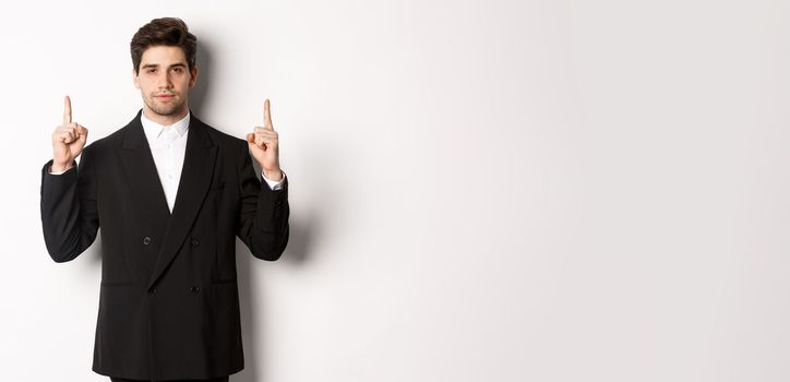 Image of confident and handsome man in formal suit, pointing fingers up, showing copy space on white background.