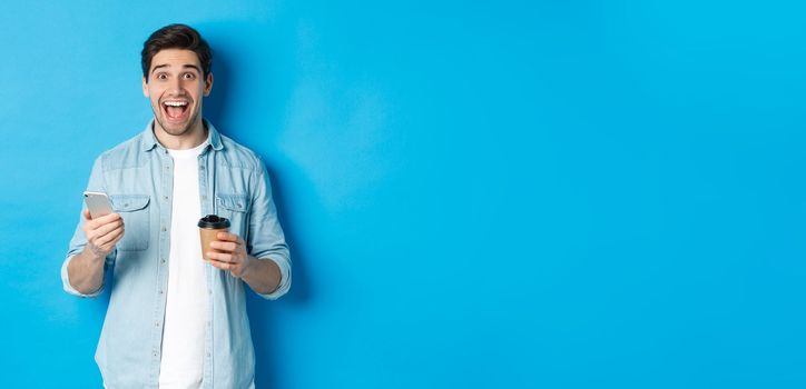 Happy young man drinking coffee and using mobile phone, looking excited, standing against blue background.