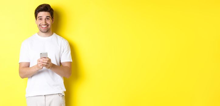 Man smiling and looking happy after reading promo offer on smartphone, standing against yellow background in white t-shirt.