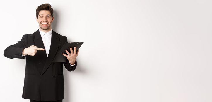 Image of handsome smiling businessman in black suit, pointing finger at clipboard with documents, standing against white background.