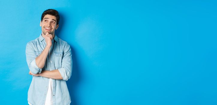 Portrait of thoughtful handsome man with beard, standing in casual outfit, looking at upper left corner and smiling, imaging or dreaming about something, standing over blue background.