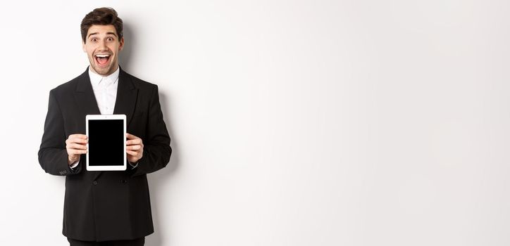 Image of attractive male entrepreneur in trendy suit, showing digital tablet screen and smiling amazed, standing over white background.