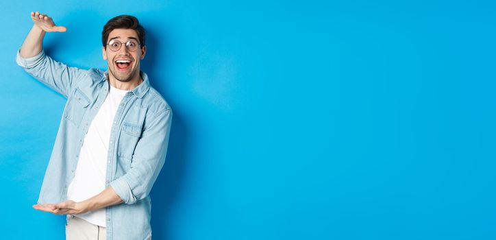 Happy man in glasses showing big size object, shaping large box, standing over blue background and smiling.