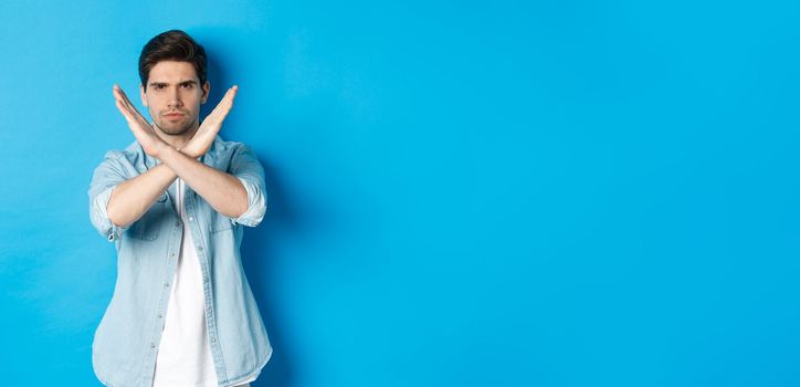 Displeased man frowning and saying no, making cross sign, tell to stop, standing against blue background.