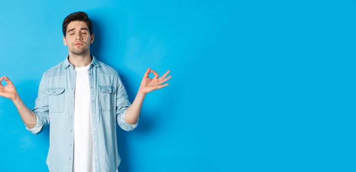 Calm man with closed eyes meditating, holding hands sideways and do yoga breathing exercises, standing against blue background.